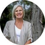 Andrea Rowe in grey cardigan and white tee shirt leaning against an ironbark tree and looking at the camera wit a smile.